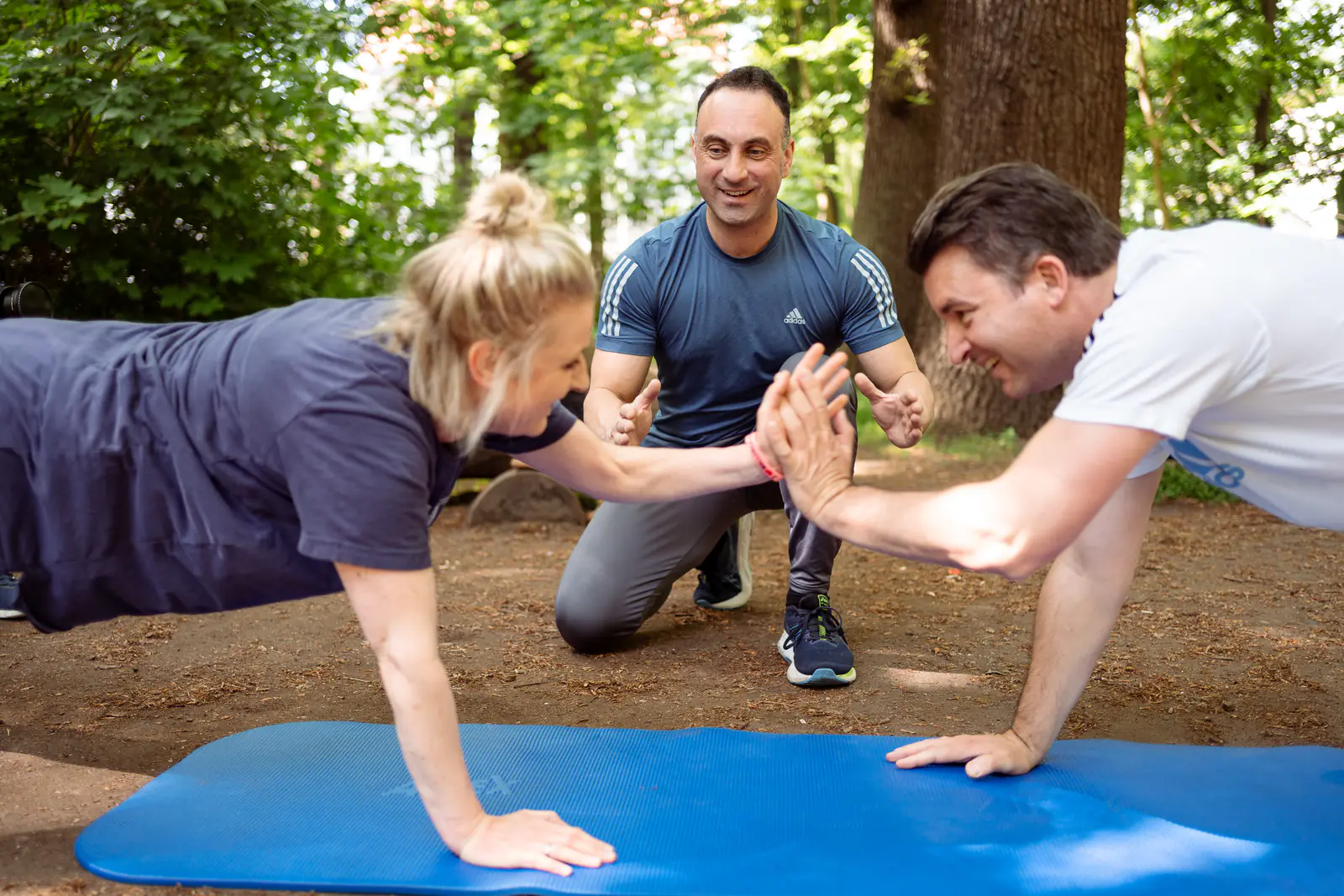Nico Fichtler kniet im Park auf dem Waldboden zwischen zwei Klienten, die sich aus der Liegestützposition auf einer blauen Matte abklatschen